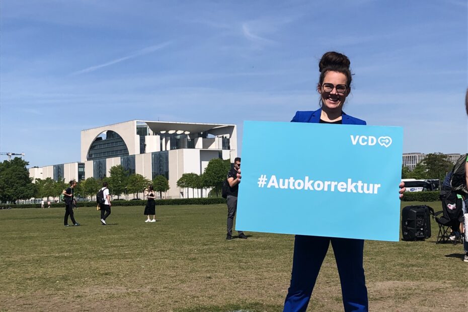 Ich stehe auf der Wiese vor dem Bundestag und halte ein blaues Schild mit Autokorrektur.