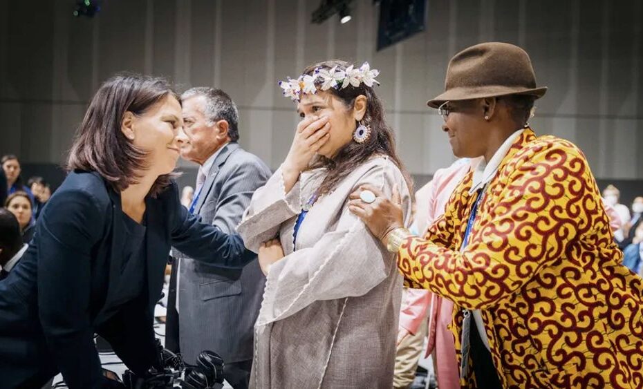 Annalena Baerbock on the left and a black person on the right are trying to comfort a woman with a wreath of flowers and a traditional-looking dress.